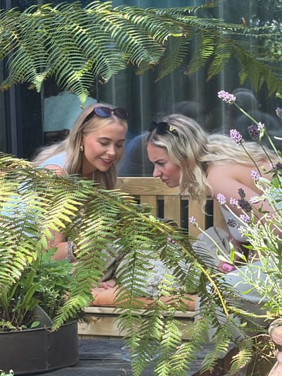 Two blonde women are partially obscured by ferns. The woman on the left wears sunglasses on her head and is smiling. The woman on the right has sunglasses on her head and is looking down. They are outside, potentially in a garden or patio setting. There are lavender sprigs present. The location cannot be ascertained to be in the city of Salford, United Kingdom.