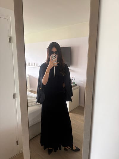 The photograph shows a woman taking a mirror selfie in a hotel room, probably in Mandelieu-la-Napoule, France. She is wearing a long black dress and a black jacket draped over her shoulders, along with black shoes. She is holding a phone in her hand and wearing sunglasses. In the background, there is a bed, a TV, and a small table with water bottles. The mirror is framed with a light wood border. The room appears to be well-lit. The flooring is light brown, likely wood or laminate.