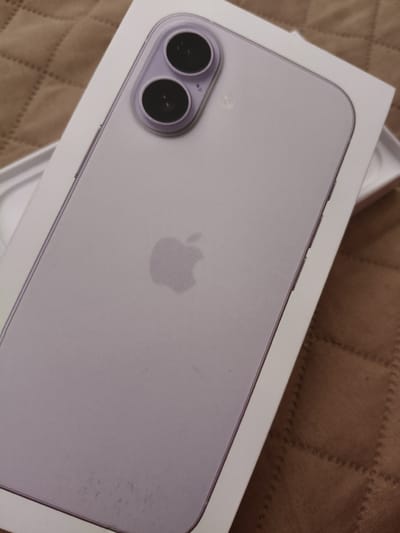 The image shows the back of a lilac-colored iPhone in its original white packaging. The phone has a dual camera system in the upper left corner and the Apple logo in the center. The background is a brown quilted material. The location in Rakitovo, Bulgaria is not evident from the image.