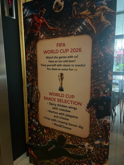 This media file is a wide photograph of a promotional poster for the FIFA World Cup 2026, featuring football players and snacks. The poster is oriented vertically.
The subjects include several stylized depictions of soccer players in action. One player in a red jersey with the number 7 is shown mid-kick. Another player in a blue jersey with the number 10 is visible. A goalkeeper in a green jersey is holding a soccer ball above their head. A fourth player, wearing a striped blue and white jersey, is also present. The background is a dynamic collage of cheering crowds, confetti, and national flags, evoking the excitement of a major football tournament.
The poster advertises "FIFA WORLD CUP 2026" and highlights a "SNACK SELECTION." Visible text details include: "FIFA WORLD CUP 2026," "Watch the games with us! Have an ice cold beer! Treat yourself with classic tv snacks! You deserve some fun 😊". Below this, under "WORLD CUP SNACK SELECTION," are listed: "• Spicy chicken wings with coleslaw," "• Nachos with jalapeno and cheese," and "• Fries with creamy bacon dip • Mac&Cheese". A golden trophy, presumably the World Cup, is also depicted.
The setting appears to be indoors, possibly a restaurant or bar, given the promotional nature of the poster and the hint of a counter or display in the foreground. The lighting suggests daytime. The overall mood conveyed by the poster is energetic and festive, focused on the enjoyment of the World Cup with food and drink. The location context provided is Budapest, Hungary.