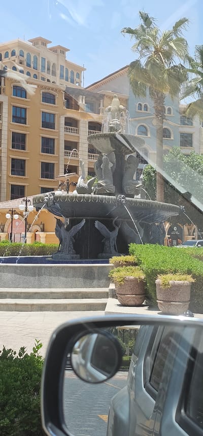 The image, taken from inside a vehicle, shows a reflection of an outdoor scene with buildings and a fountain. In the foreground, a side-view mirror of the vehicle is visible, reflecting a road. The main subject of the reflection is a large, ornate fountain featuring sculpted figures, possibly mythical creatures or animals, with water spraying from various points. Behind the fountain, there are several buildings with distinct architectural styles, featuring multiple floors, arched windows, and balconies. One building is painted in a light blue color, and another in a warm yellow or beige tone. Palm trees are also visible, suggesting a warm climate. The scene appears to be set in a public area, possibly a plaza or walkway outside a complex of buildings. There are potted plants with green foliage and some decorative urns. The lighting suggests it is daytime, with clear, bright skies. The image seems to capture a moment of travel, possibly while driving through a city, with the reflection on the car's window and mirrors providing a layered view of the surroundings. A small sign with text "LUNCH & CAKE" is partially visible at the bottom right of the reflection. The overall impression is one of an urban or resort-like environment in a sunny location.