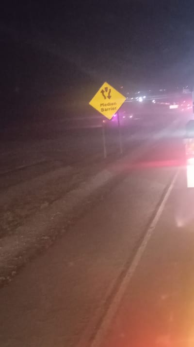 A nighttime shot reveals a "Median Barrier" warning sign, a yellow diamond with black symbols. It's positioned alongside a dark road with faint headlights visible in the background. The road surface appears wet, and the overall lighting is dim. The image has a slight reddish glow at the bottom, possibly from a vehicle's tail lights. The environment suggests a rural or suburban highway setting.