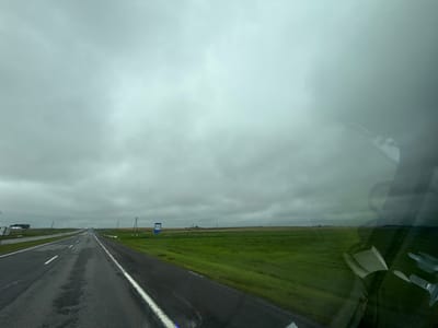 The image depicts a long, wet asphalt road stretching into the distance under a heavily overcast sky. The road has white lane markings. On the right side of the road, a vibrant green field extends to the horizon, bordered by a white fence and intermittent utility poles. A blue sign with white text is visible in the field, likely indicating a distance or place. On the left side of the road, more fields and what appear to be industrial structures are visible in the far distance. The reflection of a person and possibly the interior of a vehicle are present on the right side of the image, obscuring parts of the scene. The overall atmosphere is somber and suggestive of an uninviting, potentially rural environment in Талачынскі сельскі Савет, Belarus.