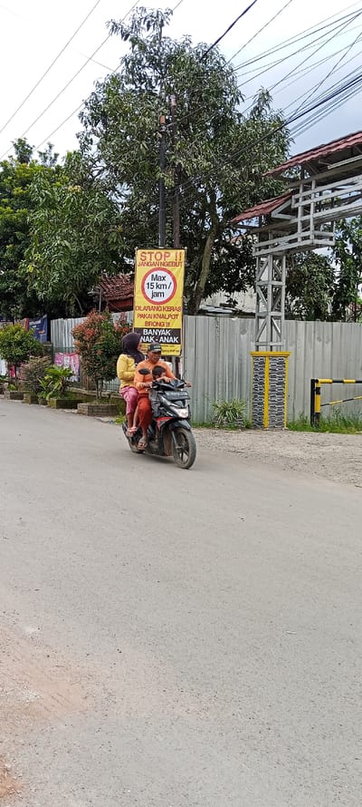 The scene takes place on a street in Cangkuang, Indonesia. The coordinates are -7.052 latitude and 107.541 longitude. Three people are riding on a motorcycle. The person driving is wearing a yellow shirt and a pink headscarf. The passenger behind them is wearing an orange shirt. A child is sitting in front of the driver. The motorcycle is black and has a license plate that reads "D 5378 CX". The motorcycle is traveling on a paved road in the direction of a sign that reads "STOP!!! JANGAN NGEBUT!!!" with a speed limit of 15 km/hr, and other text warning against loud exhausts and cautioning the presence of children. There are many electrical wires overhead. On the right side of the road is a gray fence, and behind it is a wooden structure with a red roof. There is a yellow and black metal gate in the background. There is a gray concrete sidewalk with a small patch of grass on either side of the road.