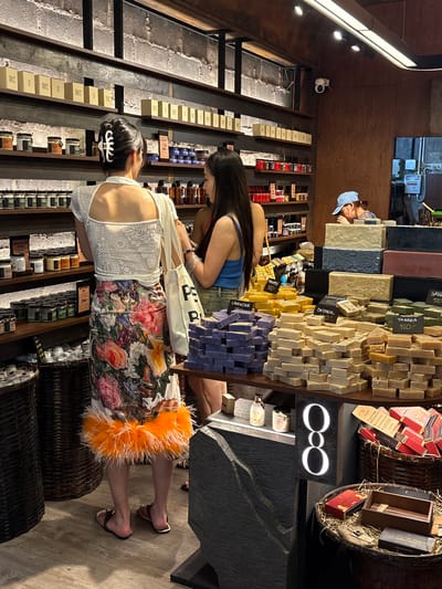 The image depicts the interior of a store in Chiang Mai City Municipality, Thailand, likely a boutique selling artisanal soaps and skincare products. Two women are browsing in the foreground. The woman on the left, with dark hair held up by a white clip, wears a white lace top and a brightly colored floral skirt with an orange feather trim. She is holding a white canvas tote bag. The woman on the right, with long dark hair, wears a blue tank top and denim shorts.
The store shelves are stocked with numerous jars and boxes of products. A prominent display of stacked bar soaps is featured on a table in the middle ground, with price tags visible for "LAVENDER 150," "PINEAPPLE 120," "OATMEAL 150," "LEMONGRASS 150," and "TANAKA 120." Another person wearing a blue cap is partially visible behind the soap display, appearing to be a staff member. The store's ambiance is rustic, with exposed brick walls and wooden shelves. In the lower right of the image, baskets filled with more products, including boxes and wooden containers, are visible. The overall lighting suggests an indoor environment.