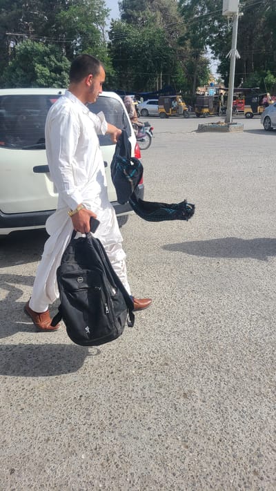 The image depicts a man standing next to a white car on a paved road in Quetta City Tehsil, Pakistan. The man is wearing a white shalwar kameez and brown shoes. He has a black backpack over his shoulder and is holding a black object in his hand. He is also holding a dark blue patterned cloth draped on the rear of the car. The background shows a street scene with trees, other vehicles, and buildings.