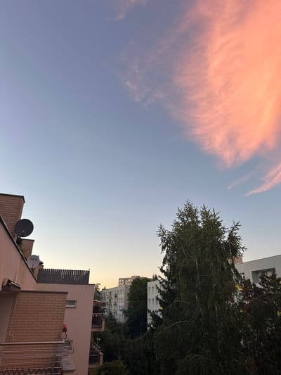 The image shows a view from an apartment balcony in Warsaw, Poland, at dusk. The sky is a gradient of blue, transitioning to a soft orange at the horizon, with wispy pink clouds illuminated by the setting sun. In the foreground, a brick building and a lighter-colored building with balconies are visible. Satellite dishes are mounted on the brick building. Further in the background, several other apartment buildings and trees are silhouetted against the sky. The overall impression is a peaceful urban scene at the end of the day.
