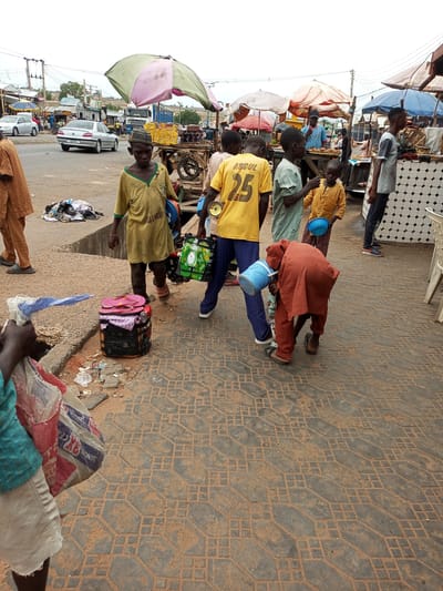 A wide shot captures a bustling street scene in Gombe, Nigeria, likely a market area based on the presence of vendors and shoppers. A young boy in a yellow t-shirt with "ABDUL 25" on the back stands near a market stall. To his left, another boy in a green shirt holds a blue bucket, and to his right, a boy in a yellow kaftan also carries a blue bucket. In the foreground, a person in an orange robe bends down, holding a blue bucket. Behind them, a few adults and children are seen in the background, some interacting with market stalls. A silver car is parked on the street, and a pile of clothing lies on the ground near the curb. The ground is paved with interlocking stones, and there are various market umbrellas and stalls scattered throughout the scene. The sky is overcast, suggesting a cloudy day.