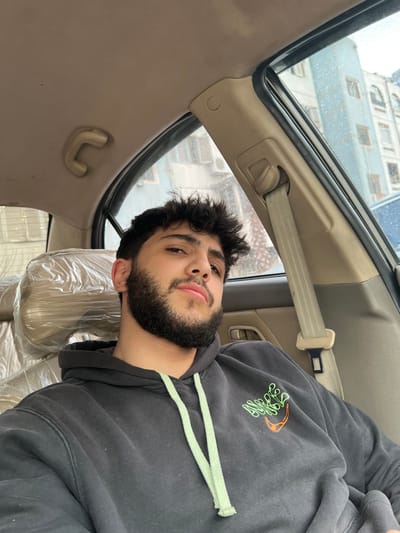 An image depicts a young adult male seated in the front passenger seat of a vehicle in Barza Municipality, Syria. The male, with dark curly hair and a full dark beard, faces forward, looking directly at the camera with a neutral expression. He is dressed in a dark grey hooded sweatshirt with light green drawstrings and a stylized green and orange graphic emblem on the left chest. The vehicle's interior features light beige upholstery and plastic trim, with a visible overhead grab handle on the left and a seatbelt assembly integrated into the right A-pillar. The passenger seat is encased in clear plastic sheeting. Through the rain-streaked passenger side window, a multi-story building with a light-colored facade, numerous windows, and external air conditioning units is visible under an overcast sky.