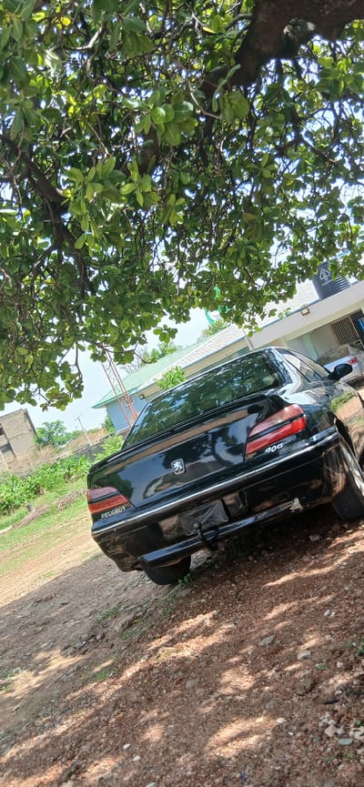 The image, originally rotated 90 degrees counter-clockwise, depicts an outdoor scene featuring the rear of a black Peugeot 406 sedan car parked on a reddish-brown dirt or gravel ground. A large, verdant tree with dense, broad green leaves and visible branches occupies the left side of the frame, casting long shadows across the ground on a bright, sunny day. The clear blue sky is visible through gaps in the foliage.
The car is seen from its rear-left quarter, displaying its distinctive red and clear taillights. The trunk lid prominently features the silver Peugeot lion emblem. The brand name "PEUGEOT" is visible in white lettering on the left taillight assembly, while the model number "406" is clearly legible on the right side of the trunk. The vehicle's surface shows signs of dust or light grime. A black tire is partially visible, resting on the unpaved surface.
In the background, a light-colored building with a distinctive greenish roof and elements of a metal structure or railing can be seen. Further back, another darker, possibly blue, structure is partially visible. No individuals are present in the image, conveying a calm and uninhabited setting. The strong sunlight, distinct shadows, and unpaved environment suggest a daytime scene in a semi-urban or rural area, consistent with the specified location of Shaba, Nigeria.