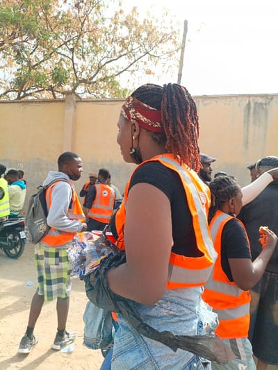 The image depicts an outdoor gathering in Sabon Gari East, Nigeria, featuring multiple individuals, many wearing orange high-visibility vests with reflective stripes, suggesting a community activity. In the foreground, a female subject is positioned with her back partially to the viewer, turned slightly to her left. She wears a black short-sleeved top, light-wash denim bottoms, an orange high-visibility vest featuring a circular logo on the back, and a patterned red and white headband. Her hair is styled in dark dreadlocks or braids, pulled back into a ponytail with reddish-brown tips. A black face mask hangs from her left ear. She holds a black plastic bag containing clear plastic items, possibly bottles or wrappers, under her left arm.

To her left, in the mid-ground, a male subject faces right, looking towards the foreground female. He wears a grey hooded sweatshirt, green and white plaid shorts, black sneakers, a black backpack, and an orange high-visibility vest. Visible on the ground near him are discarded plastic items. Further left, the rear wheel and frame of a motorcycle are partially visible. In the mid-ground to the right of the foreground female, another female subject in a black top and orange high-visibility vest is seen eating a yellow or orange food item with her right hand.

Several other individuals are visible in the background, many also in orange high-visibility vests, indicating a coordinated group. The environment includes a light-colored sandy ground, a tall, plain beige wall, and trees with sparse, light-green to brownish foliage under a bright sky.