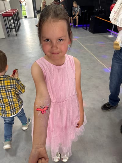 A young girl with brown hair, wearing a pink sparkly dress, stands indoors and smiles at the camera. She has a red butterfly painted on her left forearm. In the background, a boy in a yellow and black plaid shirt is visible, holding a strawberry. Other people and a black table with speakers are also present in the room, which appears to be a party or event venue. The floor is grey and smooth, with some colored light reflections. The location is not Zav'yalovo, Russia.
