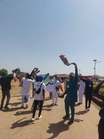 The image displays an outdoor daytime scene in Bar Gada, Nigeria, featuring approximately ten individuals on a paved road under a clear blue sky. Strong sunlight generates distinct, elongated shadows on the ground. The individuals, primarily male adults, are dressed in a mix of traditional white tunics (agbadas) and contemporary attire. Most are actively holding colorful, patterned fabric banners or flags aloft with their hands. In the foreground, center-right, a man wearing a dark teal tunic, matching pants, and brown sandals faces partially towards the camera, holding a banner overhead with his right hand; a nametag is affixed to his left chest. Behind him, another individual in a white t-shirt with "ABYIA" visible, dark pants, white sneakers, and a blue cap is seen from the back. To the far right, a solar-powered street light pole stands. The background consists of dry, lighter-colored terrain, sparse distant vegetation, and indistinct building structures beneath a hazy distant horizon line.