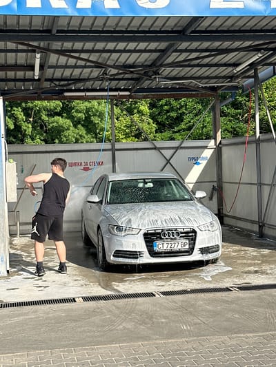 A young man in a black tank top and black shorts with white accents is washing a white Audi car at a self-service car wash. He is holding a hose and spraying the car with foam. The car is covered in a thick layer of foam. The car wash is an open-air structure with a metal roof and concrete floor. There are trees in the background. A sign above the car reads "ТУРБО ПЯНА" (Turbo Foam). Another sign on the wall behind the man reads "ЩИПКИ ЗА СТЕЛКИ" (Clips for Mats). The license plate on the car is CT 7277 BP. The scene appears to be in Kazanlak, Bulgaria, given the Cyrillic signage.