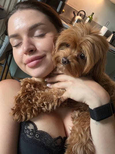 A medium shot selfie shows a young woman with closed eyes, holding a small, brown, fluffy dog. The woman is wearing a black lace bra. The dog is nestled against her chest and neck. Her right arm is visible, with a black fitness tracker on her wrist. In the background, a kitchen counter is visible with a kettle, a bottle of olive oil, and a roll of paper towels. The lighting suggests it is daytime, possibly indoors. The location is not specified.