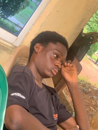 The image shows a young Black male sitting in an outdoor setting in Ukukwa, Nigeria. He is wearing a black t-shirt with white text that reads "geared protection". His right hand is raised to his face, with his fingers touching his eye. The background consists of a yellow-painted wall and a glimpse of greenery, suggesting a warm climate. The man is seated in a light-colored plastic chair. The overall scene appears casual and relaxed, with the man perhaps tired or thoughtful.
