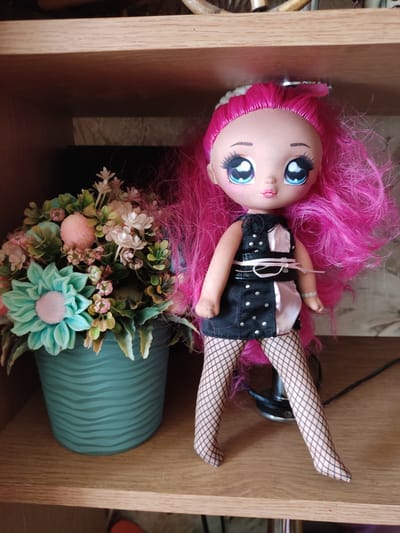 A doll with bright pink hair stands on a wooden shelf next to a pot of artificial flowers. The doll is wearing a black dress with silver accents and fishnet stockings. The pot of flowers contains a variety of artificial blooms in shades of green, pink, and white, with a large teal flower as the centerpiece. The background shows the interior of a room with wooden shelving and a dark wall. There is no visible indication of location within Kryvyi Rih, Ukraine.