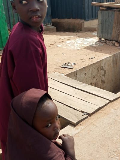 In Tempure, Nigeria, an outdoor scene unfolds with two young individuals. The foreground is dominated by a toddler, whose face is partially obscured by a dark brown head covering. The toddler is looking to their right, their expression a mixture of curiosity and apprehension. To the left and behind the toddler stands a slightly older child, also looking to their right, with a visible portion of their face showing. This child wears a maroon garment.
The environment is rustic, characterized by sandy ground. A concrete structure, possibly a well or drainage area, is present in the mid-ground. A wooden plank bridge, made of rough-hewn planks, spans over the concrete structure, providing a pathway. In the background, a dark, corrugated metal structure is visible, suggestive of a building or enclosure. Scattered debris, including what appear to be discarded flip-flops, litter the sandy area. The lighting indicates it is daytime, with clear shadows cast by the objects and individuals.