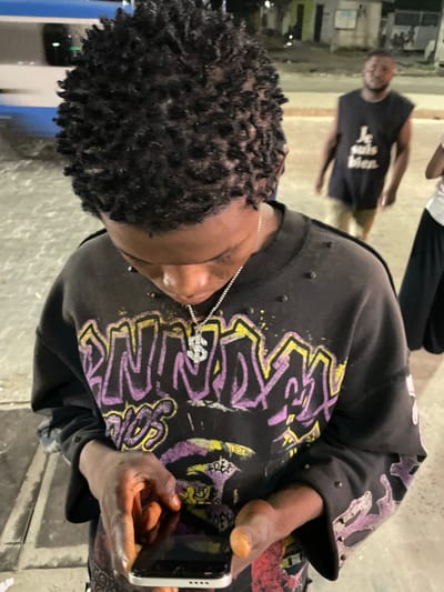 A medium shot captures a young Black male from the chest up, looking down at a smartphone in his hands. He has short, dark, coiled hair and is wearing a black crewneck sweatshirt adorned with a vibrant, graffiti-style graphic in purple and yellow. A silver chain with a dollar sign pendant rests on his chest. The background is dimly lit, suggesting an outdoor urban setting at night. In the mid-ground, to the right of the primary subject, another Black male stands facing away from the camera, wearing a black sleeveless t-shirt with white text that reads "Je suis bien." To the far right, partially visible, is a third individual wearing a white top and black pants. The environment appears to be paved with concrete, and a blurred blue and white object, possibly a vehicle or bus, is visible in the far left background. The overall impression is a candid street scene in Ikorodu, Nigeria.