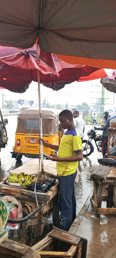 The image captures a street scene in what appears to be Maiburiji, Nigeria, based on the visible features. A young man, dressed in a yellow shirt with blue sleeves and dark pants, stands beside a wooden cart laden with bananas and some goods in black plastic bags. He is looking down at his phone. The cart is under a partially torn, multi-layered sunshade. In the background, there is a yellow "keke," a type of auto-rickshaw common in Nigeria. Further back, people can be seen walking, and a motorcycle is parked nearby. The road and pavement appear wet, suggesting it has recently rained. Wooden stools or crates are visible at the edge of the frame. Overall, the image depicts a typical scene of informal commerce and transportation in a Nigerian urban area.