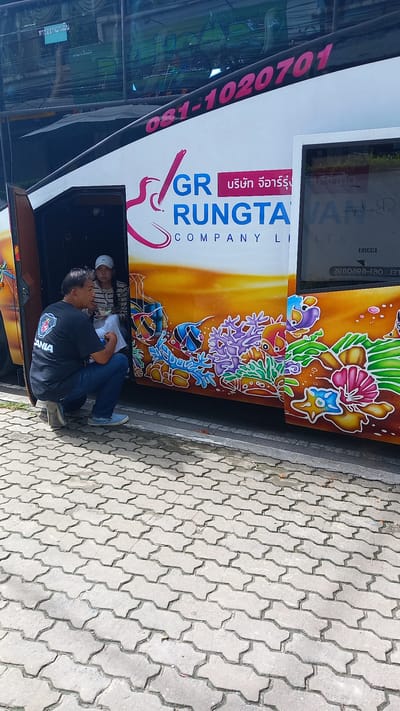The image captures a scene in Ban Ao Nang, Thailand, showing a man wearing a black Scania t-shirt kneeling near the open door of a bus. Another person is visible inside the bus. The bus has "GR Rungta Company Limited" printed on its side, along with a phone number and decorative graphics. The ground is paved with interlocking concrete blocks.