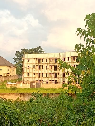 This media file captures an outdoor scene in Ukukwa, Nigeria, featuring a multi-story building under construction. The building, partially covered by scaffolding made of wooden poles and planks, has multiple windows and appears to be several stories high. To the left of the construction site is a smaller, completed building with a dark, sloped roof and light-colored walls.
In the foreground, lush green vegetation, including trees and bushes, dominates the lower portion of the image, partially obscuring the view of the buildings and suggesting a natural or semi-urban environment. A low, light-colored wall runs across the middle ground, separating the dense vegetation from the buildings and the grassy area surrounding them.
The sky above is a pale, hazy blue with hints of clouds, suggesting it might be late afternoon or early morning, with soft lighting. The overall atmosphere is calm and serene, with no specific activity or event clearly depicted beyond the ongoing construction. There is no visible text in the image.