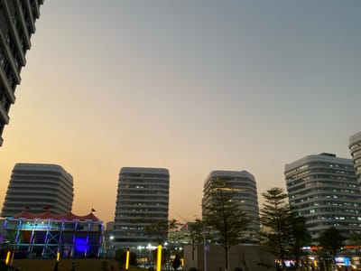 The image depicts an urban scene in Guang Zhou Shi, China, captured during dusk or twilight hours. The sky transitions from a warm, golden-orange near the horizon to a lighter blue-grey overhead, indicating the time of day.

The scene is dominated by several modern, high-rise buildings featuring distinctive rounded corners and horizontal window bands. One building is partially visible on the far left, while four additional similar structures are prominent in the mid-ground and background, with internal lights illuminated.

In the lower-left quadrant, a large, multi-level temporary structure stands out. It has a tent-like roof with sections of red and purple fabric and is brightly illuminated by vibrant blue and purple lights, suggesting an ongoing event, fair, or festival. Scattered trees and other low vegetation are visible in the foreground and between the buildings.

On the ground level, several indistinct figures of people are present, some near the colorful temporary structure and others further to the right, implying a public gathering or evening stroll. Yellow ground-level pole lights or fixtures also contribute to the artificial illumination of the area. No discernible text is visible on any signs or structures within the image. The overall atmosphere suggests a lively evening in a modern city.