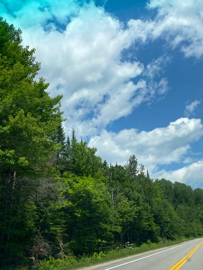 The image shows a paved road with yellow double lines, curving to the right, bordered by a dense forest of green trees on the left. The sky above is blue with scattered white cumulus clouds, and a turquoise hue in the upper left corner. The trees are lush and appear to be in full summer foliage. The terrain slopes upwards from the road into the forest, with rocky outcrops visible in places. The scene appears to be a natural, rural setting, likely a highway or scenic route. There are no people or structures visible in the image, suggesting a remote or undeveloped area. The image was taken in the city of Lac-Simon, Canada.