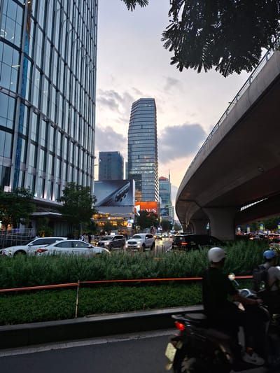 This image depicts an urban scene in Thủ Đức, Vietnam, likely during the evening or dusk, as indicated by the artificial lighting and the soft, diffused natural light. The perspective is unusual, with the camera tilted at an angle, making it appear as if the viewer is looking up at the cityscape.
The foreground is dominated by a modern glass-fronted building, its facade reflecting the surrounding environment and creating a grid-like pattern. To the right of this building, a street runs parallel to a lush green hedge. Traffic is visible on the street, with cars and motorcycles forming a queue, suggesting moderate congestion. Several people are on motorcycles, some wearing helmets.
In the background, a distinctive skyscraper with a curved, segmented facade stands tall, illuminated by internal lights. Below it, a large billboard displays a smartphone, possibly an advertisement for Samsung. The overall setting is a busy urban environment with a mix of contemporary architecture and infrastructure, including what appears to be an elevated road or bridge structure arching overhead. The sky is cloudy, with hues of orange and grey suggesting the transition from day to night. The scene is alive with activity, characteristic of a bustling city. Visible text includes "Samsung" and "HAUS".