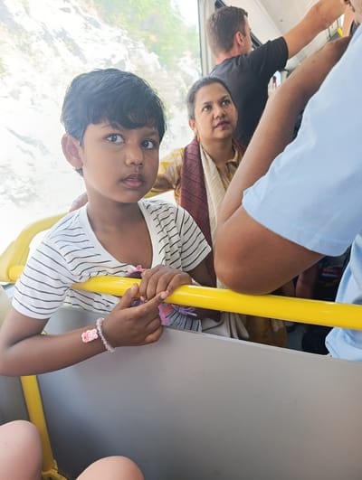 The image shows a group of people on a bus. In the foreground, a young girl with dark hair and brown eyes looks directly at the camera. She is wearing a striped t-shirt and a pink beaded bracelet with a bear charm on her wrist. Her hands are resting on a yellow handrail. Behind her, a woman with dark hair and a light-colored scarf looks to her left. Further back, a man in a black t-shirt is visible, holding onto a vertical pole. The setting appears to be a bus with large windows, suggesting an urban or scenic route. The lighting indicates it is daytime. The overall mood is casual and observational. No visible text is present in the image. The location context is Dražin Vrt, Montenegro.