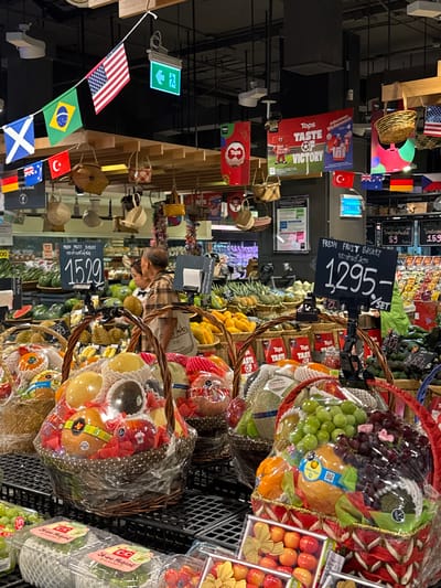 The image is a photograph taken at a Tops supermarket in Chiang Mai City Municipality, Thailand. The scene is focused on a display of fresh fruit baskets for sale. In the foreground, several fruit baskets are prominently featured, wrapped in plastic. One basket, on the right, contains a variety of fruits including green and red grapes, a large yellow pear, and avocados. Another basket, on the left, holds different fruits, including what appear to be pears and possibly a melon. In the middle ground, two people, an older man and a woman, are standing and looking at fruit. The man is wearing a plaid shirt, and the woman is wearing a striped top. Behind them, more fruit and vegetables are displayed on shelves. The background shows the interior of the supermarket, including overhead lighting and signage. There are also flags hanging from the ceiling, including the American flag, the Brazilian flag, and the Scottish flag, which are part of a decorative display. Pricing tags are visible, with one indicating "1295-" and another "1529". The overall impression is a vibrant and well-stocked produce section of a supermarket.