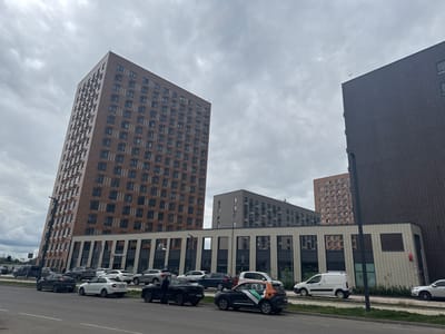 The media file depicts an outdoor scene in Shcherbinka, Russia, under an overcast sky. The composition is dominated by modern residential buildings with distinct architectural styles. A tall, slender building with a brick facade and numerous windows stands prominently on the left, tilting slightly. To its right, a shorter, rectangular building with a gray facade and repeating window patterns is visible. Further to the right, a taller, dark-colored building occupies the edge of the frame. In front of these buildings, a low-rise commercial structure with large windows and a striped facade runs horizontally.

Parked cars line the street in front of the buildings, including a white van, a black SUV, a white sedan, and other vehicles. A few individuals are visible near the parked cars, one person appears to be standing next to a black SUV, and another is walking on the sidewalk. The scene suggests a typical urban environment with residential and commercial spaces. The weather appears cloudy, typical for a temperate climate.

A red circular logo with white text, possibly a store or company sign, is visible on the side of the commercial building. The overall impression is one of a modern, developing urban area. No specific event or activity is clearly taking place beyond normal daily life.