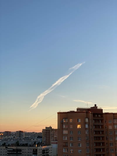 The image captures a wide-angle view of a city skyline at sunset, with a prominent apartment building in the foreground. The sky is a gradient of blue, transitioning to orange and yellow near the horizon. A wispy contrail stretches diagonally across the upper portion of the sky, with a small dark object, likely an aircraft, visible near the contrail.

The foreground features a multi-story brick apartment building with numerous windows. Several windows on the building reflect the warm hues of the sunset, creating glowing rectangles of light. The roof of this building is visible, topped with antennas and a small structure.

Behind the foreground building, a dense cityscape unfolds, composed of various apartment buildings of different heights and architectural styles. These buildings are also illuminated by the sunset. Power lines are strung across the scene, connecting different structures. The overall impression is of a residential area in a Russian city at dusk. The presence of identical apartment blocks suggests a Soviet-era housing development, common in cities like Sinyavino, Russia, known for its Soviet heritage and subsequent urban development.