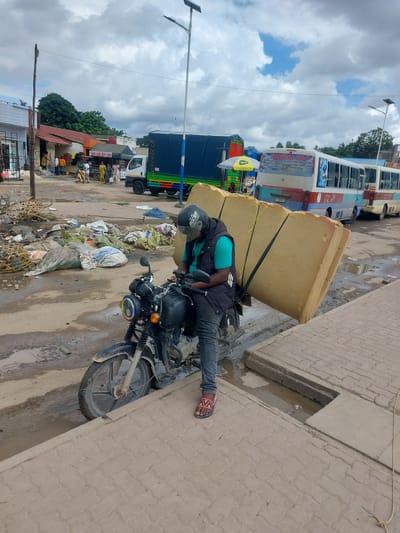 The image shows a man riding a motorcycle with a large foam mattress strapped to the back in Dar es Salaam, Tanzania. He is wearing a helmet, a green t-shirt, a black vest, and sandals. The motorcycle is black and appears to be struggling to support the oversized load. The setting is an urban area with a mix of parked vehicles, including a bus, a truck, and other vehicles partially visible. The street is littered with trash and debris. Pedestrian pathways are paved with bricks and are also stained with water and grime. Buildings and shops are visible in the background, along with utility poles. The sky is overcast with scattered clouds.