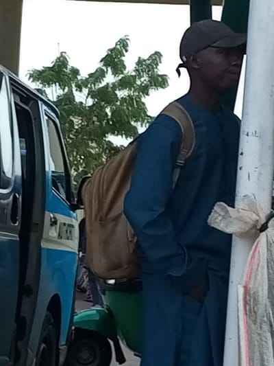 A full-body shot shows a young man leaning against a white pole, his right shoulder and cheek pressed against it. He is wearing a dark blue long-sleeved shirt and matching pants, and a dark gray baseball cap that casts a shadow over his face. A tan backpack is slung over his left shoulder. To the left of the man, a blue and white vehicle, likely a commercial bus or taxi, is parked with its side door open. The vehicle has a white stripe with black lettering running along its side, and part of its front is visible. Behind the man and the vehicle, a lush green tree with many branches and leaves is visible against an overcast sky. In the foreground, at the base of the white pole, a white plastic bag is tied around it, with a portion of a pink and white striped bag visible to its right. The scene appears to be on a street in Gombe, Nigeria, possibly near a bus stop or market.