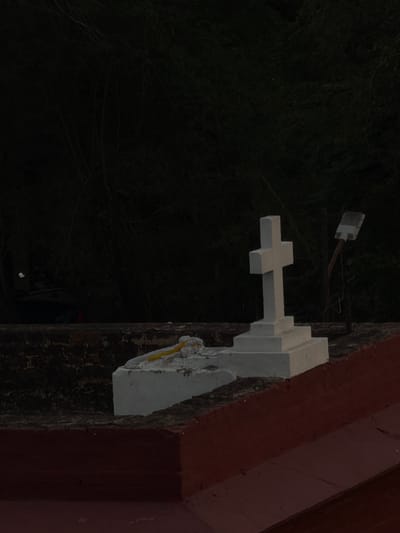 The image shows a white cross tombstone with a tiered base, situated on a raised platform. To the left of the cross, a smaller white structure is visible, adorned with what appears to be yellow and white flowers. In the background, dark foliage suggests a wooded or overgrown area, while the foreground features a reddish-brown surface, possibly part of a roof or wall. A metal pole with a light fixture extends upwards to the right of the tombstone. The lighting suggests it is either dawn or dusk, with low visibility and a somber atmosphere. The scene is consistent with a graveyard or memorial site within Delhi, India.