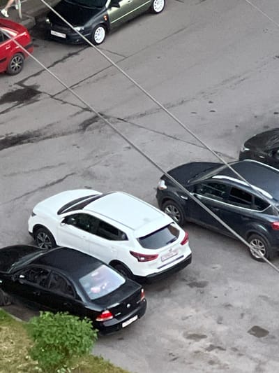 The image shows a street scene in Sinyavino, Russia, with several parked cars and overhead wires. In the foreground, a black sedan is parked on the left, next to a patch of green bushes. To its right, a white crossover SUV is parked. Behind the white SUV, a dark blue SUV is parked. Further back and to the left, a red car is parked. In the upper left corner, the rear of a green car is visible, with a person standing nearby wearing white sneakers and dark pants. The ground appears to be asphalt with some wet patches and tire marks. The overhead wires are a prominent feature, crisscrossing the scene.
