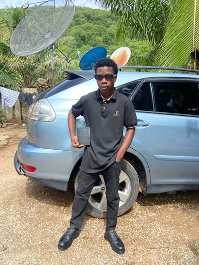 A young Black male stands in front of a light blue SUV parked on a gravel surface. He is wearing a black polo shirt with a colorful "GIVENCHY" logo on the left chest, black pants, black sunglasses, and black dress shoes. He holds a black book with "HOLY BIBLE" printed in gold on its cover in his right hand. His left hand is in his pocket. Behind him, there are several satellite dishes mounted on poles, some palm trees, laundry hanging on a line, and lush green hills in the background. The setting appears to be a residential outdoor area in Mararaba, Nigeria.