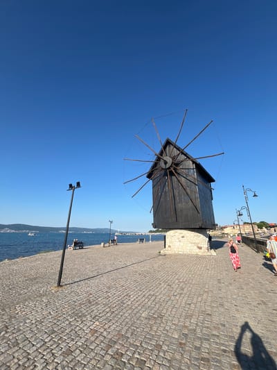 This media file depicts a daytime scene in Nesebar, Bulgaria. The primary subjects include a traditional black wooden windmill with its sails visible against a clear blue sky. In the background, a cobblestone promenade runs alongside a body of water, likely the Black Sea.

Several people are present on the promenade. One person is wearing a red and white patterned dress, and another is seen from behind with a striped shirt and brown bag. Further in the background, more individuals are scattered along the waterfront and near buildings. Objects of note include several lampposts lining the promenade, benches where people are seated, and some boats visible on the water.

The setting is a picturesque coastal town with a historic feel, suggested by the architecture and the windmill. The bright sunlight and clear sky indicate fair weather and a time of day likely late morning or early afternoon. The overall emotion conveyed is one of leisure and pleasant outdoor activity. There is no visible text in the media file.