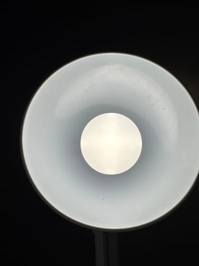 This is a close-up, overhead shot of a modern white lamp with a dark background. The lamp shade is a wide, shallow cone, and at its center is a bright, circular light source, likely an LED bulb. The inside of the shade appears slightly textured and has a few small, dark smudges. The overall impression is one of simplicity and focused illumination. The image does not contain any elements that can be used to determine its location in Bietigheim-Bissingen, Germany.