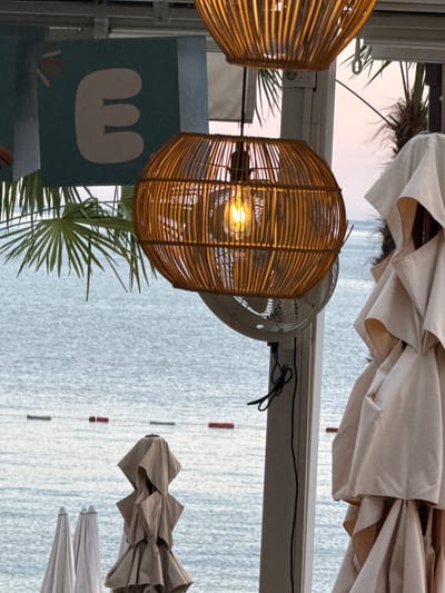 The image depicts a seaside scene in Kamenovo, Montenegro, likely at a beach cafe or resort. In the foreground, several closed beach umbrellas are stacked, with one prominently featured in beige fabric on the right side. Above them, a woven rattan lamp with a lit bulb hangs, casting a warm glow. Behind the umbrellas and lamp, the calm, blue sea stretches towards the horizon. A line of red and green buoys marks a boundary in the water. Palm fronds are visible in the upper left, and a partial view of a sign with a stylized letter "E" on a teal background is also present. The sky above is a soft pink and orange, indicating either sunrise or sunset. A white ceiling fan is partially visible beneath the rattan lamp.