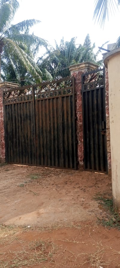 The image displays the exterior of a property entrance in Alor, Nigeria, featuring a metal gate structure. The scene is dominated by a weathered dark corrugated metal double gate, flanked on the right by a narrower, similar pedestrian gate equipped with a latch mechanism. Both gates feature decorative, ornate scrollwork and square patterns along their upper edges. These gates are integrated into reddish-brown pillars and wall sections, textured with a repeating circular or patterned relief design. The tops of the pillars are flat and weathered. To the far right, a light-colored, possibly concrete, cylindrical structure is partially visible. The ground directly in front of the gates consists of reddish-brown dirt or sandy soil, with scattered patches of dry, sparse green and brown vegetation. The background reveals dense green foliage, predominantly palm trees, beneath a bright, overcast sky. No individuals or active subjects are present in the frame.