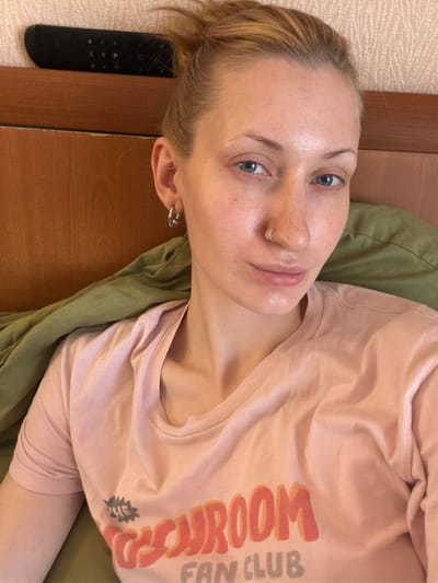 A close-up, overhead shot shows a young woman with blonde hair tied up, wearing a light pink t-shirt with orange and yellow text that reads "ROOM FAN CLUB." She has blue eyes, light skin, a nose ring, and a hoop earring in her left ear. She is lying down on a green blanket or pillow, and behind her is a wooden headboard. A black remote control is visible on top of the headboard. The lighting suggests it is daytime, and the scene appears to be indoors, likely a bedroom. There is no information to determine her location in the city of Unknown, Russia.