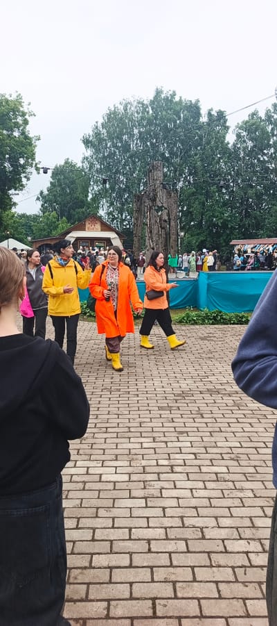 The image depicts an outdoor scene with a brick pathway in the foreground. Several people are walking on the pathway. On the left, a person wearing a black hooded jacket and dark pants is seen from behind. Moving towards the center, three women are walking abreast. The woman on the left wears a yellow jacket and dark pants. The central woman is in a bright orange raincoat and dark pants, holding a dark bottle. The woman on the right is wearing an orange jacket and dark pants. Behind them, a large, carved wooden sculpture of a person stands prominently. Further in the background, a wooden building with signage, crowds of people, and trees are visible, suggesting a public gathering or event. The overall atmosphere appears to be cloudy.