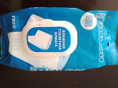 The image shows a package of wet toilet paper. The package is blue and white with Russian text. The text includes "ВЛАЖНАЯ ТУАЛЕТНАЯ БУМАГА" which translates to "wet toilet paper" and "ALCOHOL FREE". There is also a circular logo with "MON" and "МЯГКИЙ УХОД" which translates to "soft care". The package is lying on a dark, flat surface, likely a table. There is no indication of the location being Irkutsk, Russia, or any other specific geographical area. There are no people, actions, or interactions visible in the image.