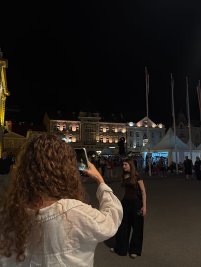 In Novi Sad, Serbia, at night, a person with curly brown hair, wearing a white shirt, holds up a smartphone to take a picture of another person. This second person, a woman with long brown hair, is wearing a dark brown off-the-shoulder top and wide-leg black pants. She is holding a clear wine glass in her right hand. In the background, illuminated buildings with traditional architecture and flags are visible. Several white tents are set up, suggesting an outdoor event or market. People are milling about in the background, and a statue of a person is visible between two buildings. The scene appears to be a public square at night.