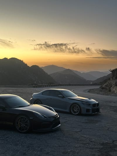 Two sleek cars parked on a mountain road at sunset. The first car is black with yellow accents and the second is a light gray color. Rolling mountains fading into the background under a hazy sunset sky. The road winds through the mountains. The image is rotated 90 degrees counter-clockwise.