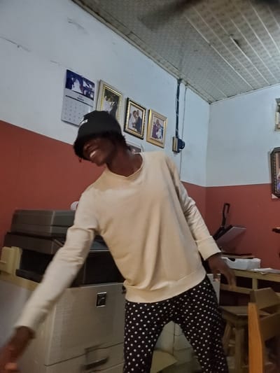 An indoor scene in Mararaba, Nigeria, features a dark-skinned individual positioned centrally, smiling with teeth visible and head tilted left. The person wears a black bucket hat, a light-colored long-sleeved top, and black pants with white polka dots. The left arm is extended towards the lower left, while the right hand rests near wooden furniture on the lower right.

The room's walls are bifurcated horizontally, with the lower section painted reddish-brown and the upper section white. The ceiling consists of light-grey, possibly corrugated, panels.

On the left side of the frame, a large, light-grey office multi-function machine (likely a copier/printer) with a silver emblem is visible. Above it on the white wall, a calendar for "MARCH" displays a wedding image, flanked by multiple framed photographs, some with gold frames, depicting couples.

Towards the right side, another framed photograph hangs on the white wall. A dark, possibly electrical, conduit or strap descends vertically along the corner where the wall colors meet. Further right, a partial view of a wooden stool or chair is present, along with what appears to be a heat press or similar device on a surface. The overall environment suggests an office or a domestic space with office equipment.