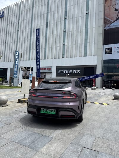In the Huangpu District of China, a purple Xiaomi SU7 electric vehicle is parked on a paved plaza in front of a modern building with a vertical facade. The car has a green license plate that reads "A B73955". Signs for "MOEN" and "SHIBA" are visible on the building, as well as a large banner for "FOTILE". The sky is overcast, and the lighting suggests it is daytime.