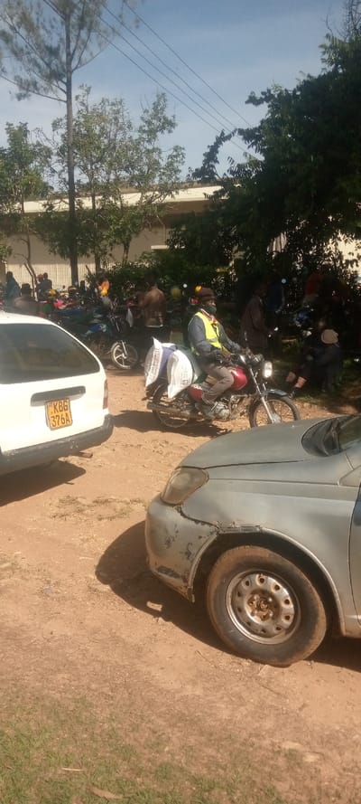The image shows a dirt road in Malava, Kenya, with several vehicles parked on either side.  A white car with a "KBG 376A" license plate is parked on the left side of the road, and a gray car is parked on the right. Several motorcycles are parked behind the cars. A man wearing a yellow safety vest is riding a motorcycle, and he has a large bag on the back.  The road is surrounded by trees and greenery. The sky is clear and blue.